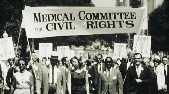 Large multiracial group of men and women holding signs and marching on a street towards viewer.