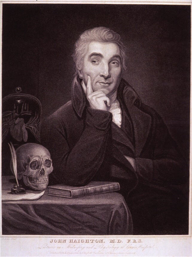 A man (John Haighton) sitting and looking to the left by a table with a book and skull on it.