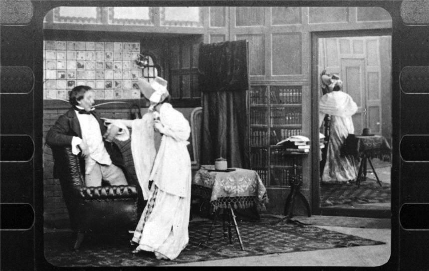 Woman in hat and white dress stands beside a man kneeling on a leather chair in a room with mirror.