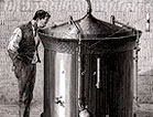 A man stands next to a large cylindrical beer vat and gazes into the vat through a small opening in the conical cover.