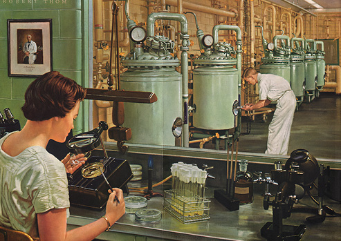 A woman seated at a laboratory bench examines a petri dish under a magnifying glass.  In the background a man examines an industrial fermentation tank.