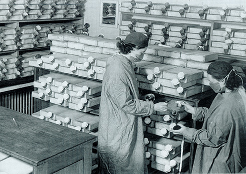 Two women in surgical gowns, masks, and head coverings in a room stacked with square ceramic penicillin vessels.