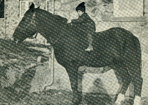 Black and white photograph of a child on the bare back of a dark colored horse in front of a stone building.