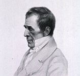 Man with dark hair and eyebrows in a jacket sitting in profile facing left.