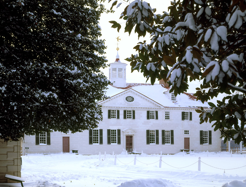 Large white house with many black shuttered windows on a snow covered landscape with trees.