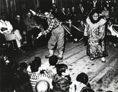 An Eskimo man and an Eskimo woman dance while several men beat drums; many people look on, children sit in the foreground.