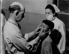 Dr. W. P. Whitted examines the eyes of a young Native American trachoma patient, Trachoma School, Fort Defiance, Arizona, 1941. A female nurse wearing a mask stands behind the patient. Courtesy National Archives and Records Administration.
