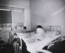 Two women in three-bed ward Indian Sanitorium, Albuquerque New Mexico 1951. One woman is sitting on the middle bed while the other is sitting in a chair eating food next to the third bed.