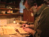 An image of Mohamed Zakariya at his workbench writing on a piece of paper. His left hand is holding down the paper and he is holding a quill in his right hand.