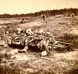 Sepia-toned stereograph of decaying corpses on a battleground. Three men are gathering the remains for burial.
