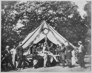 Fotografía en blanco y negro de un grupo de hombres uniformados y un cirujano preparándose para realizar una amputación, frente a una carpa del hospital.