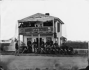 Black and white photoraph of a large group of uniformed solders standing in front, and on the balcony, of a white, two-story building.