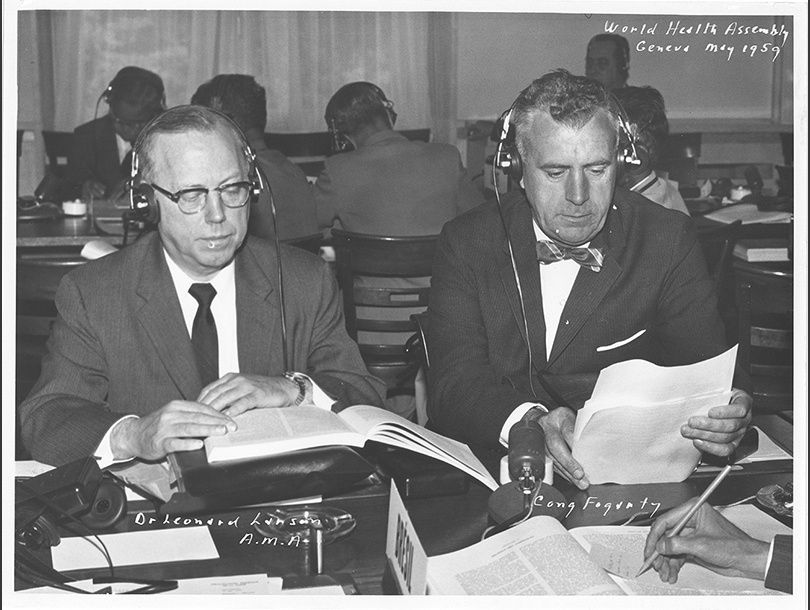 Two white men seated together reading while wearing headphones.
