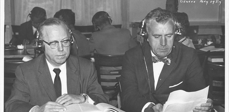 Two white men seated together reading while wearing headphones.