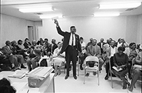 An African American man stands amongst a crowd of seated observers holding up a check.