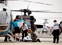 Uniformed workers help a patient laying on a gurney into a helicopter, other helicopters standby in the background.