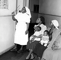 An African nurse stands leading a class with African mothers and their children seated.