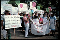 A group of Latino men and women stand together on a dirt path with signs, banners, and balloons.