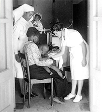 A white woman leans to assess an African child on his mother’s lap, while an African nurse holding another child watches on the other side of the mother.