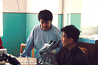A female doctor, standing, talks to a man to her right who is facing a microscope.