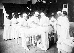 Three surgeons standing in an operating room performing surgery with several nurses and attendants observing.