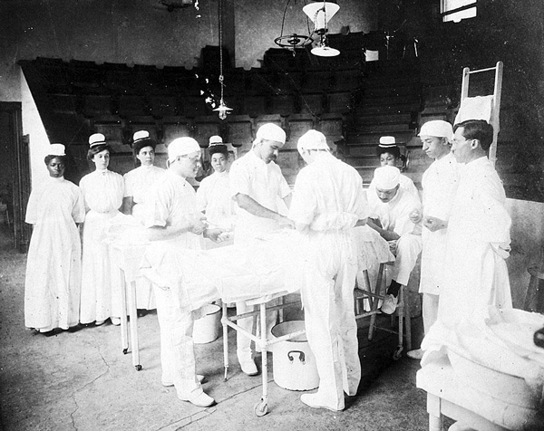Three surgeons standing in an operating room performing surgery with several nurses and attendants observing.