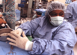 Surgeon in scrubs, mask, gloves and cap seated at an operating table.
