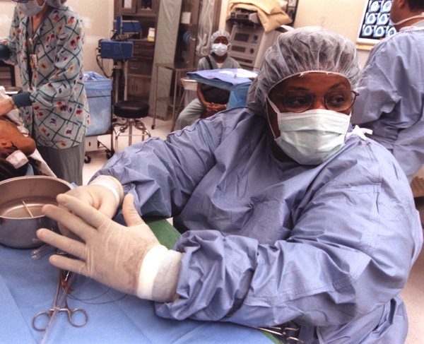 Surgeon in scrubs, mask, gloves and cap seated at an operating table.