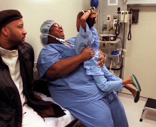 Two people sit while one in scrubs holds up a child.