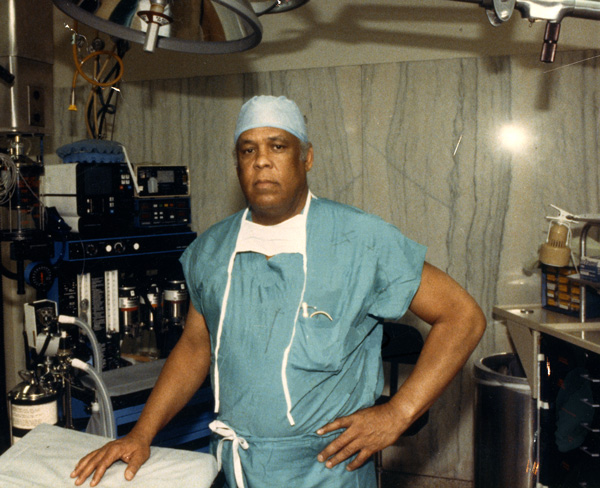 Man in green surgical scrubs and head covering standing in an operating room.
