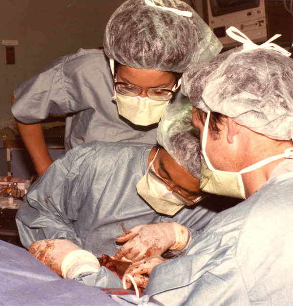 Three in surgical scrubs and masks in surgery: two next to the patient and one looking over a surgeon into the operation. 