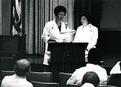 A woman in white medical coat standing at a lectern.