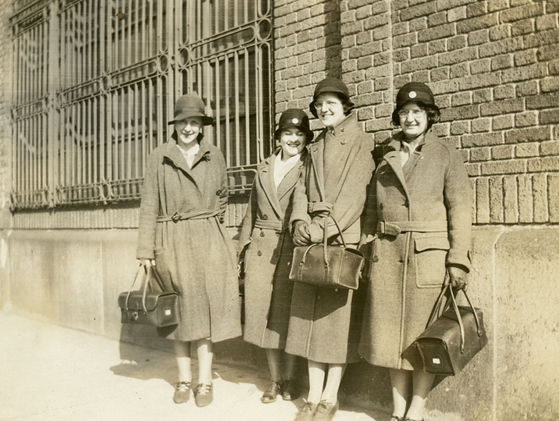 Four White female nurses in overcoats and cloche hats holding leather nursing bags and standing next to a brick building.
