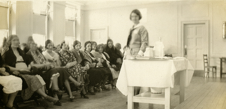 A uniformed, White, female nurse standing next to a table with an audience of seated women in front of her.
