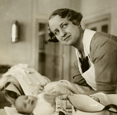 White, female nurse bathing infant on a table, metal pitchers and small glass jars in front.