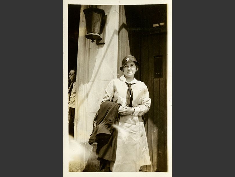 White, female nurse in cloche hat standing in a doorway, overcoat over arm.