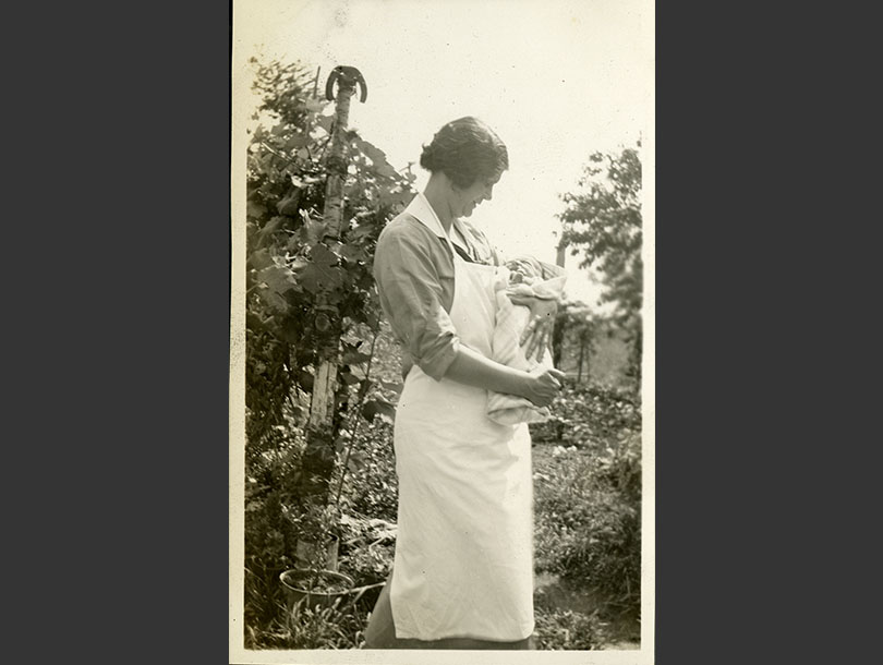 White, female nurse in a white apron holding a swaddled infant in a garden.