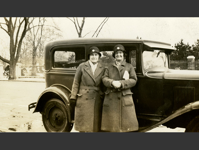 Two White female nurses in matching winter overcoats and cloche hats standing next to a Ford Victoria.