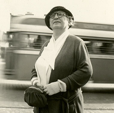 Middle-age White woman wearing a hat, glasses, and gloves, standing on a street with a bus passing behind her.