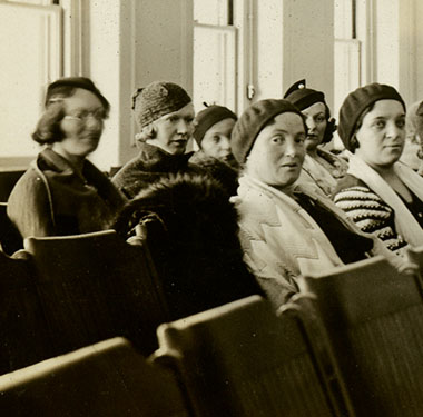 Three rows of women sitting in coats and hats in front of a table.