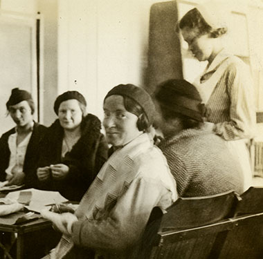 Five mothers seated around a table with supplies and two White female nurses helping with project.
