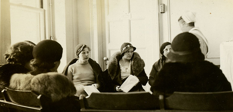 Sitting in a semi-circle, 7 females in coats watch a White female nurse in uniform conducting class.