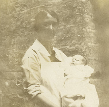 White female nurse in white apron holding an infant outside.