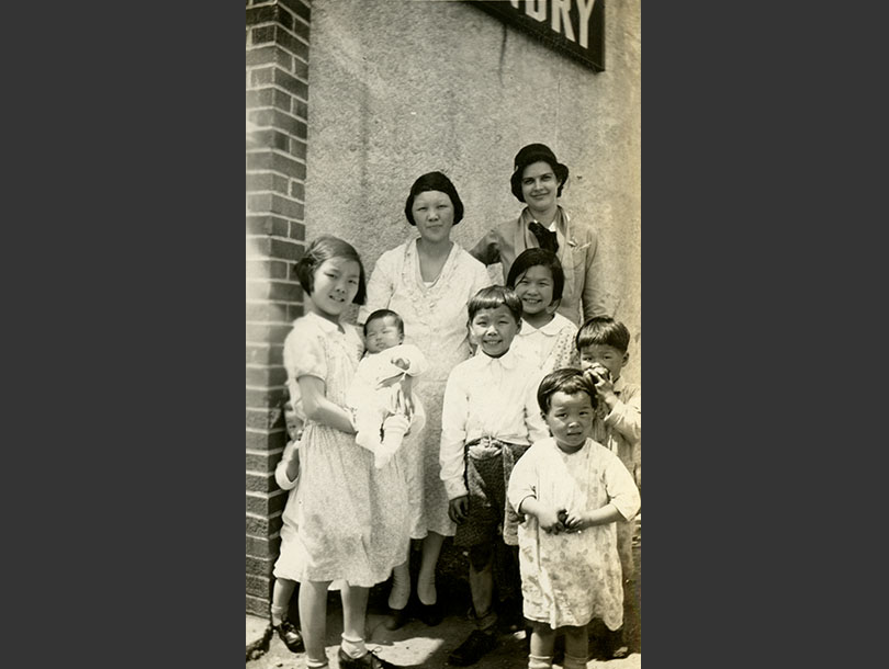 White female nurse in uniform posing with a Chinese mother and six of her children outside a laundry.