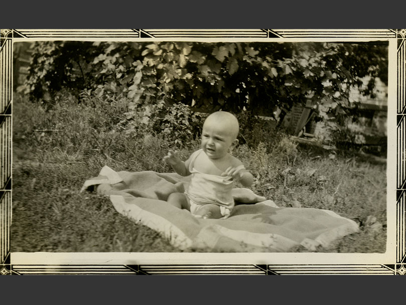 A White baby sitting up on a blanket in a grassy spot in the sun.
