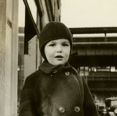 White toddler boy in winter coat and hat standing on a Bronx Street, elevated train line in background.