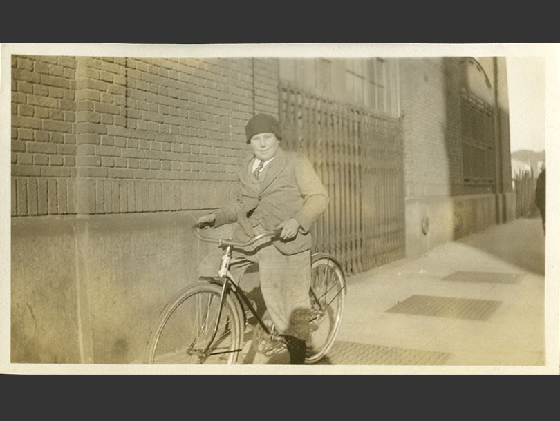 Boy posing with his bicycle on a sidewalk outside a brick building in Gun Hill.
