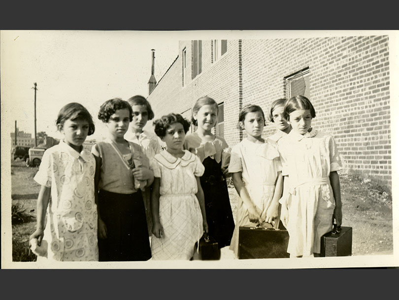 8 school girls in dresses, some carrying small cases, on their way to a day camp.
