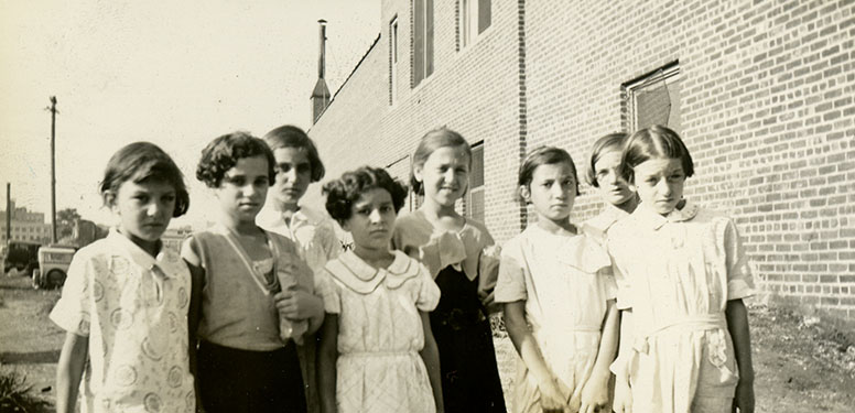 8 school girls in dresses, some carrying small cases, on their way to a day camp.