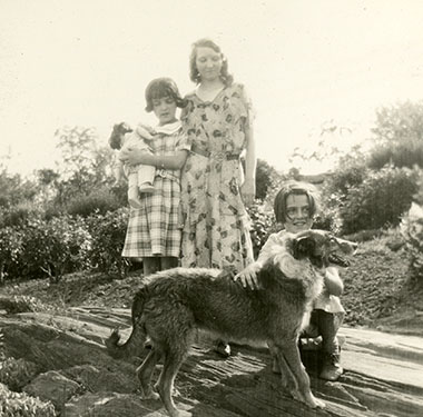 Three sisters in dresses with a dog outside on a rock, shrubs in background.